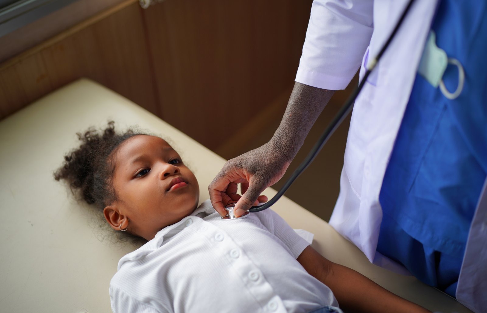 A clinician listens to a young girl's chest with a stethoscope as she lies on an examination table
