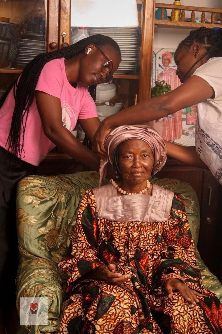 Volunteers help an older woman tie her headscarf as part of personal care.