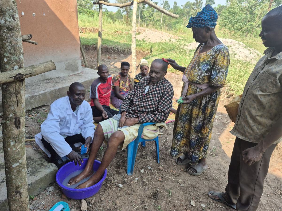 A community health worker washes a patient's feet while a family caregiver combs his hair.